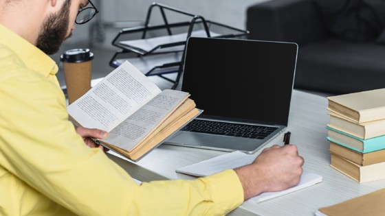 Man som läser en bok vid sin laptop. Foto: Mostphotos