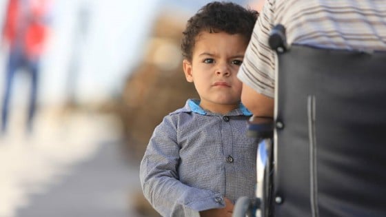 Litet barn som står vid sidan om sin far i rullstol