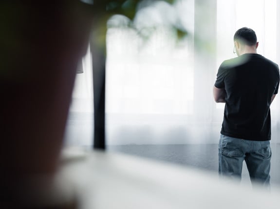 Bild tagen bakifrån på en man som står med armarna i kors vid ett fönster. Foto: Mostphotos