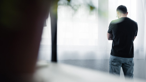 Bild tagen bakifrån på en man som står med armarna i kors vid ett fönster. Foto: Mostphotos