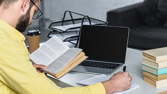 Man som läser en bok vid sin laptop. Foto: Mostphotos