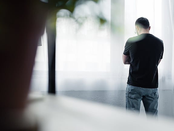 Bild tagen bakifrån på en man som står med armarna i kors vid ett fönster. Foto: Mostphotos