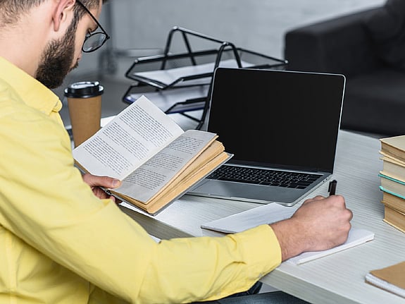 Man som läser en bok vid sin laptop. Foto: Mostphotos