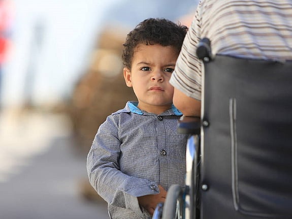Litet barn som står vid sidan av sin far i rullstol