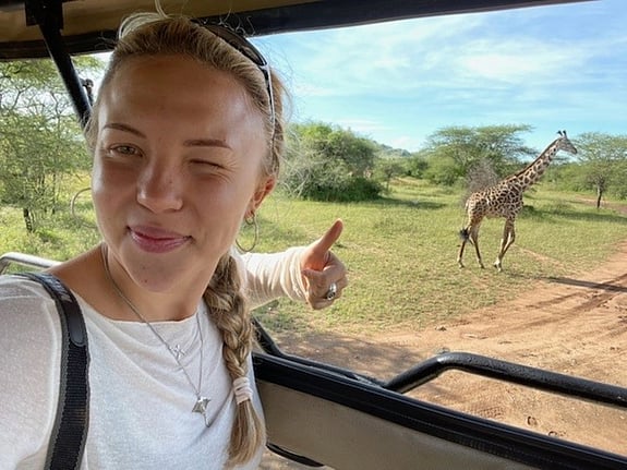 Linnea sitter i en bil och tar en selfie med en giraffe i bakgrunden. Foto: Privat